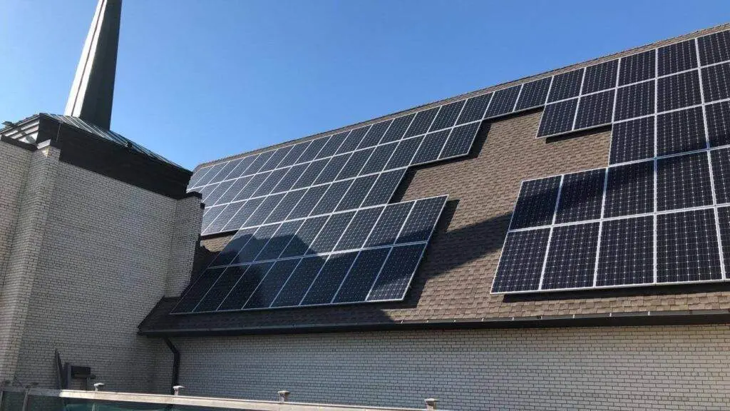 Numerous solar panels are installed on the brown shingled roof of a light brick church, adjacent to a steeple, showcasing a sustainable energy solution like those supported by the Xcel Solar Battery Storage Program.